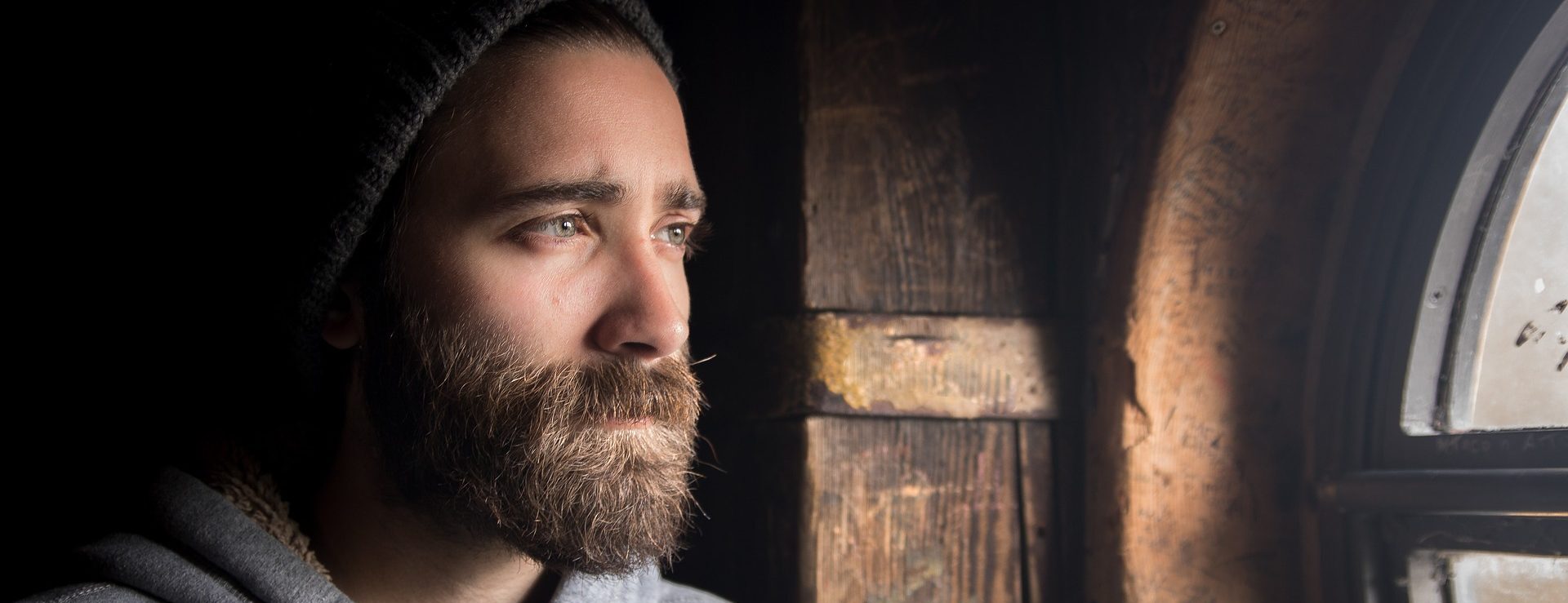 Ein bärtiger Mann mit einer Beanie-Mütze blickt mit ernster Miene aus dem Fenster und steht neben einer Holzwand.