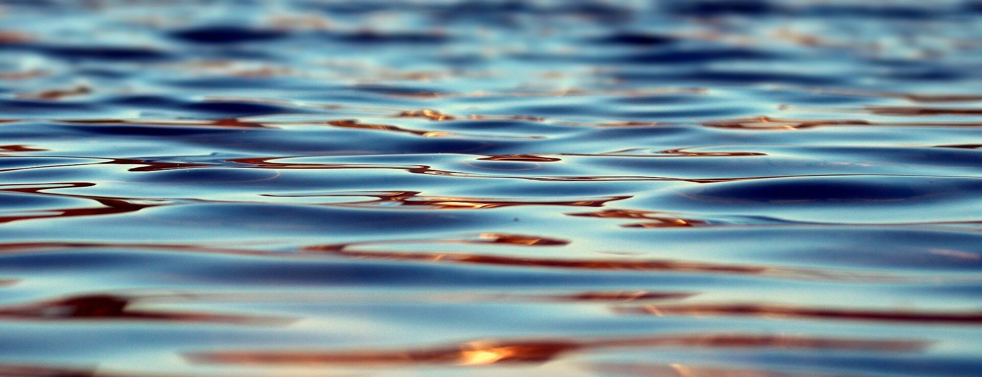 Nahaufnahme sanfter Wellen auf der Oberfläche eines Gewässers, die das Sonnenlicht in Blau- und Orangetönen reflektieren.