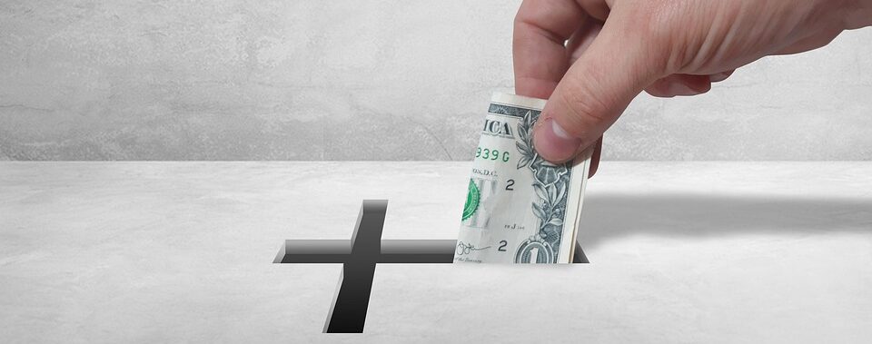 Eine Hand steckt einen gerollten US-Dollarschein in einen Schlitz mit einem kreuzförmigen Ausschnitt auf einer grauen Oberfläche.