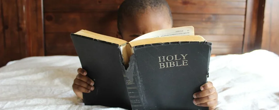 Ein Kind liest in einer großen abgenutzten Bibel, im Hintergrund eine Holzpaneelwand.