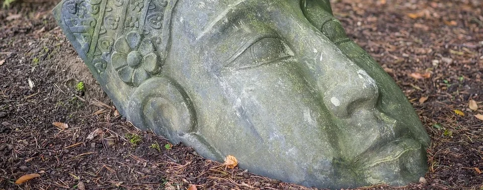 Steinskulptur eines großen menschlichen Kopfes, teilweise im Boden vergraben, umgeben von Erde und gefallenen Blättern.