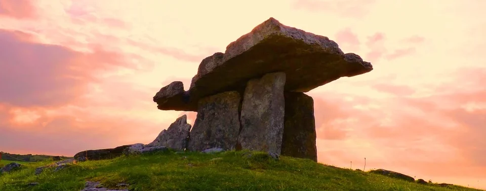 Eine prähistorische Steinstruktur erhebt sich vor einem leuchtenden Sonnenuntergangshimmel und ist von Grasland umgeben.
