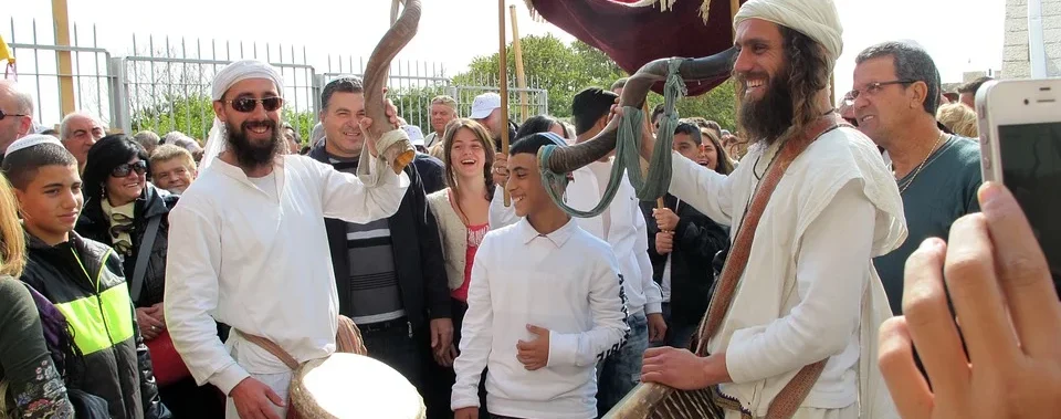 Eine Gruppe von Menschen versammelt im Freien, darunter einige Männer in weißen Gewändern und Kopfbedeckungen, die ein großes gedrehtes Hornblasinstrument halten.