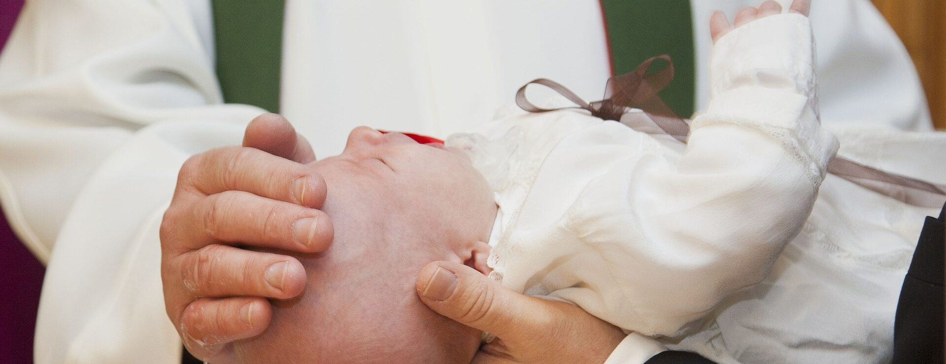Ein Baby in weißer Kleidung wird während einer religiösen Taufzeremonie von einem Prister in einem weißen Gewand gehalten.