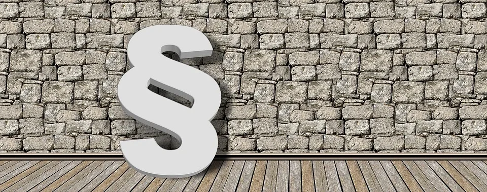 Großes weißes Pragraphensymbol, das an einer Steinmauer lehnt, im Vordergrund ist ein Holzboden zu sehen.