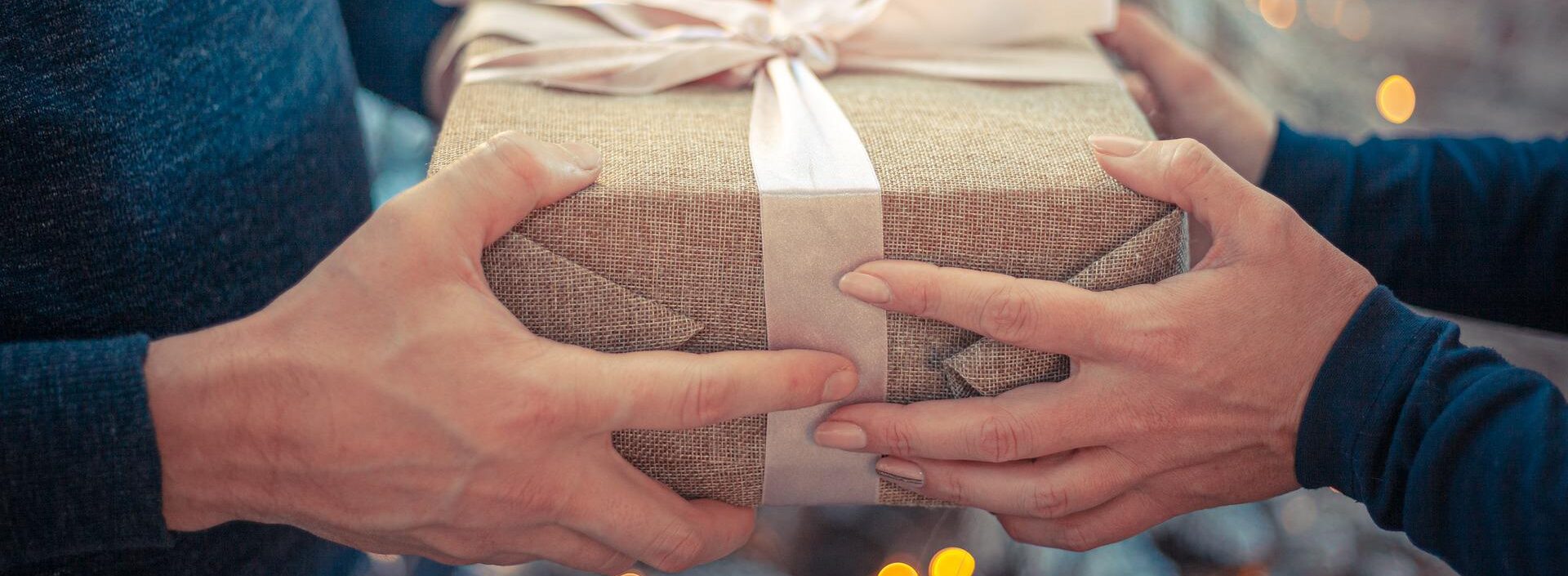 Zwei Personen halten eingewickeltes Geschenk mit einer Sackleinen Struktur und einer cremefarbenen Schleife, im Hintergrund sind festliche Lichter zu sehen.