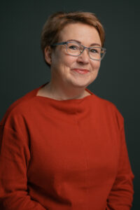Claudia Flieder: kurze Haare; Brille; roter Oberteil, posiert vor einem dunklen Hintergrund.