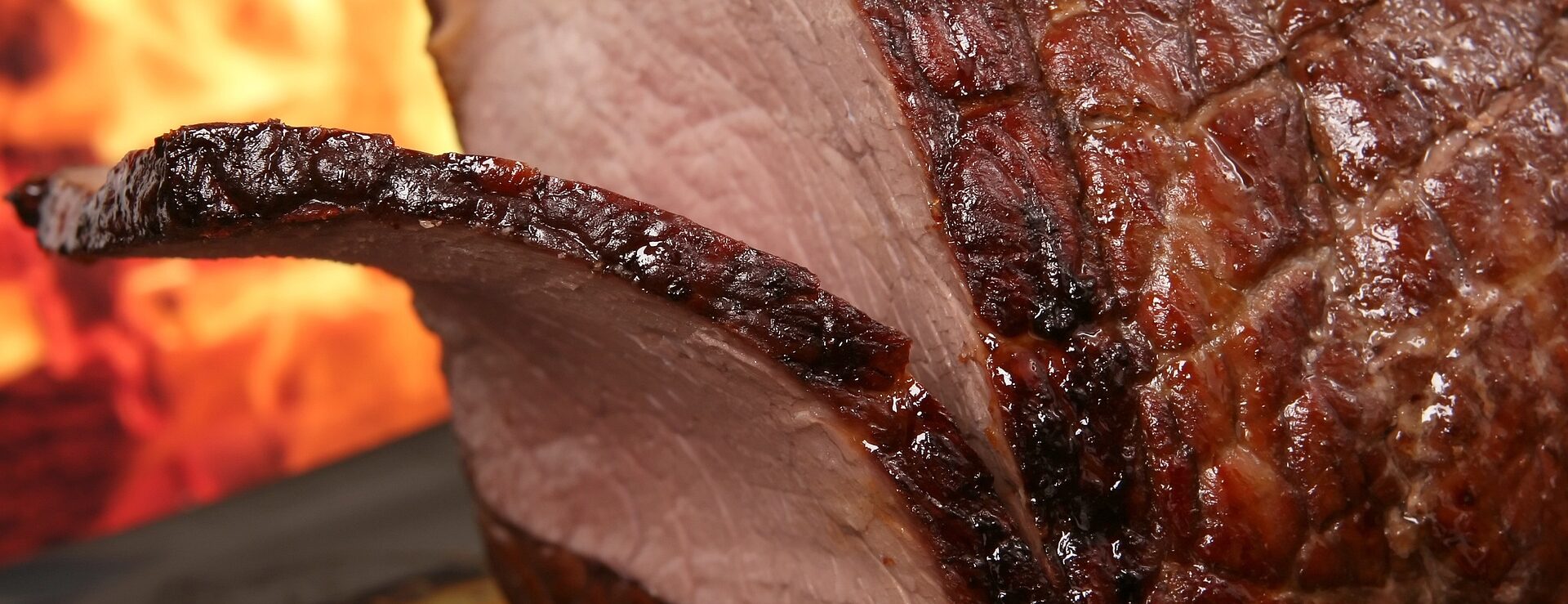 Nahaufnahme eines geschnittenen, saftigen Bratens mit knuspriger Kruste, im Hintergrund ein Feuer.