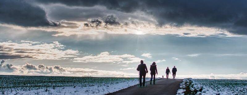 Menschen gehen auf einer Landstraße mit Schnee an den Seiten unter einem dramatisch bewölkten Himmel mit Sonnenstrahlen.