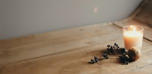 Brennende Kerze auf einem Holztisch mit dunklen Beeren und einem kleinen Dekorationsobjekt vor einer grauen Wand.