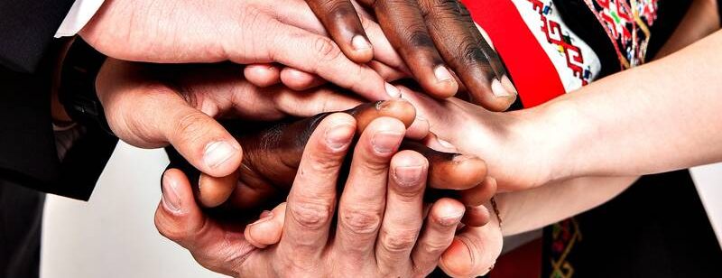 Verschiedene Hände verschiedener Hautfarbe übereinander symbolisieren Einheit und Teamwork.