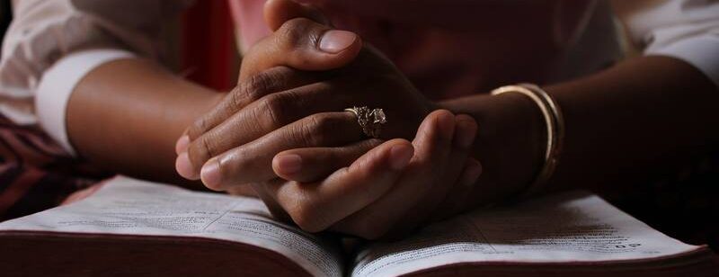 Eine geöffnete Bibel liegt auf einem Tische und darauf liegen gefaltete Hände