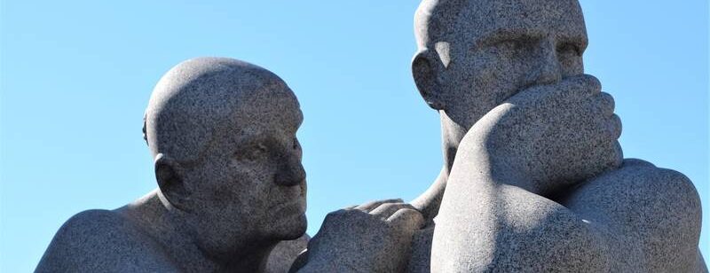 Zwei Steinskulpturen: ein Mann mit der Hand vor dem Mund, ein anderer beugt sich nah heran, unter einem klaren blauen Himmel.