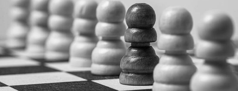 Nahaufnahme von Schachfiguren, die auf einem Schachbrett aufgereiht sind, mit einer dunklen Figur zwischen hellen Figuren.